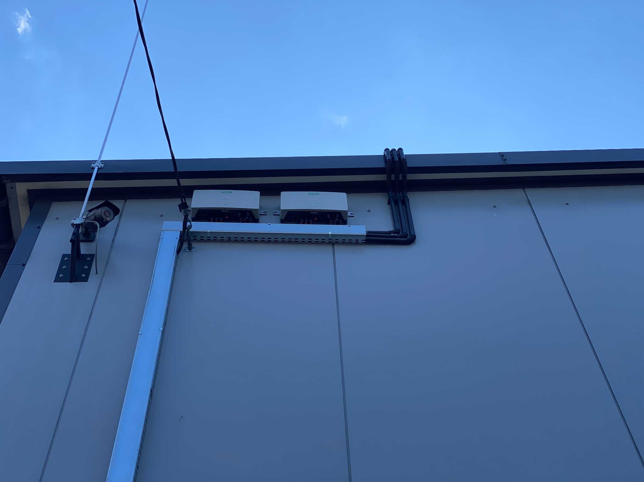 Dwa falowniki fotowoltaiczne zamontowane na elewacji budynku, widoczne czarne rury kablowe, kamera przemysłowa i maszt odgromowy na tle błękitnego nieba.