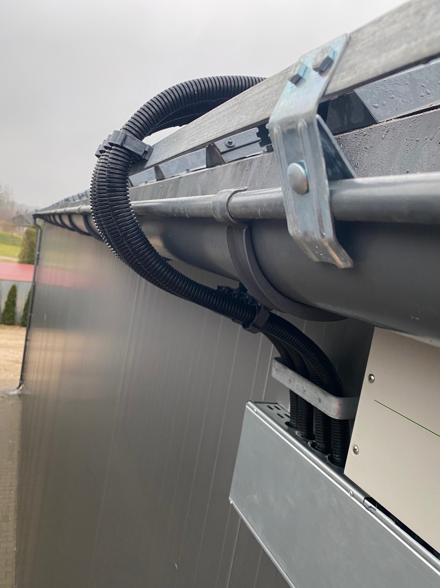 Kabel ochronny prowadzony wzdłuż rynny do urządzenia, mocowany metalowymi uchwytami, widok z dołu na szarym tle budynku.