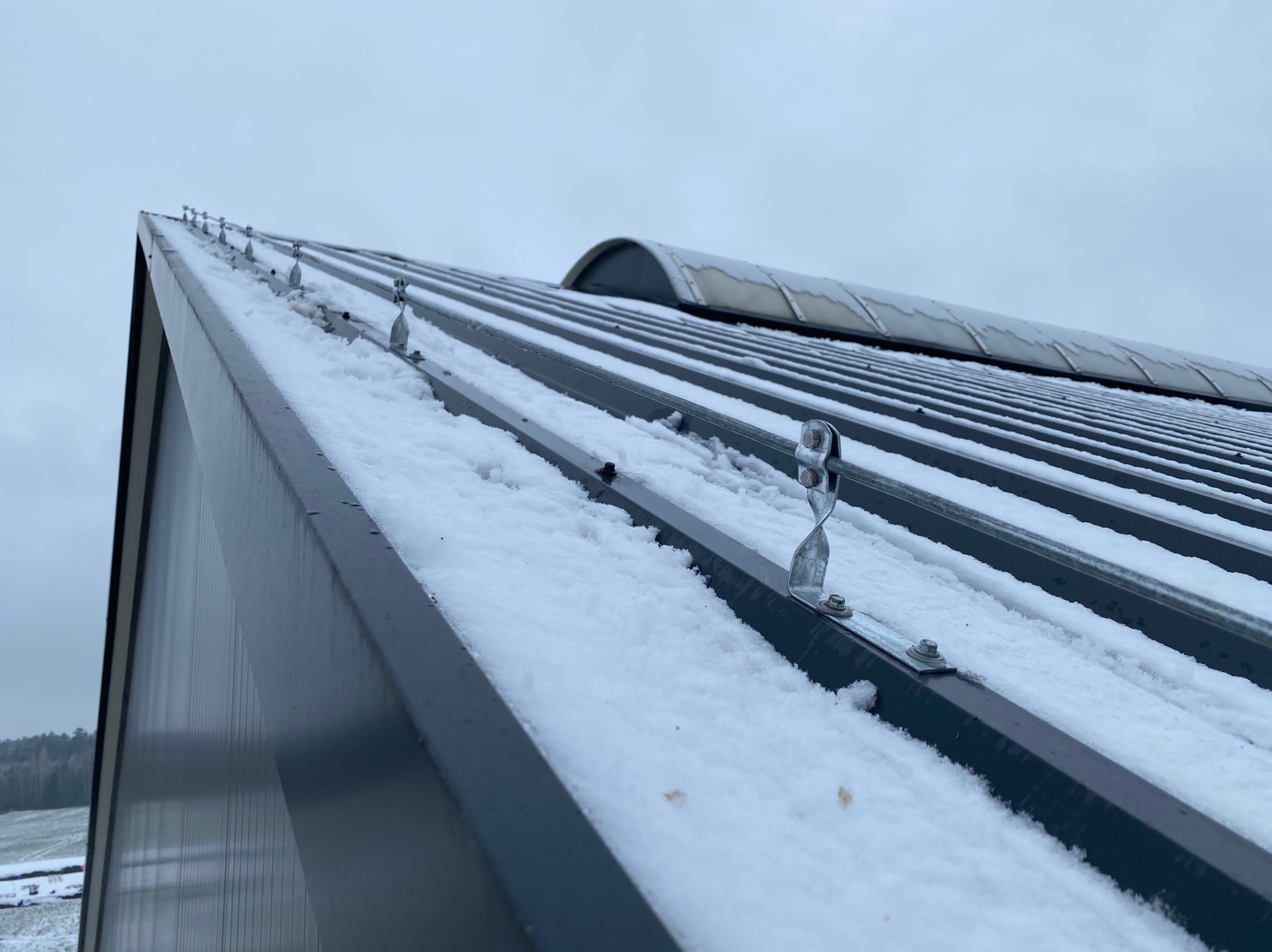Metalowy dach pokryty śniegiem z widocznymi uchwytami montażowymi i elementami konstrukcyjnymi, w tle zalesiony teren i pochmurne niebo.