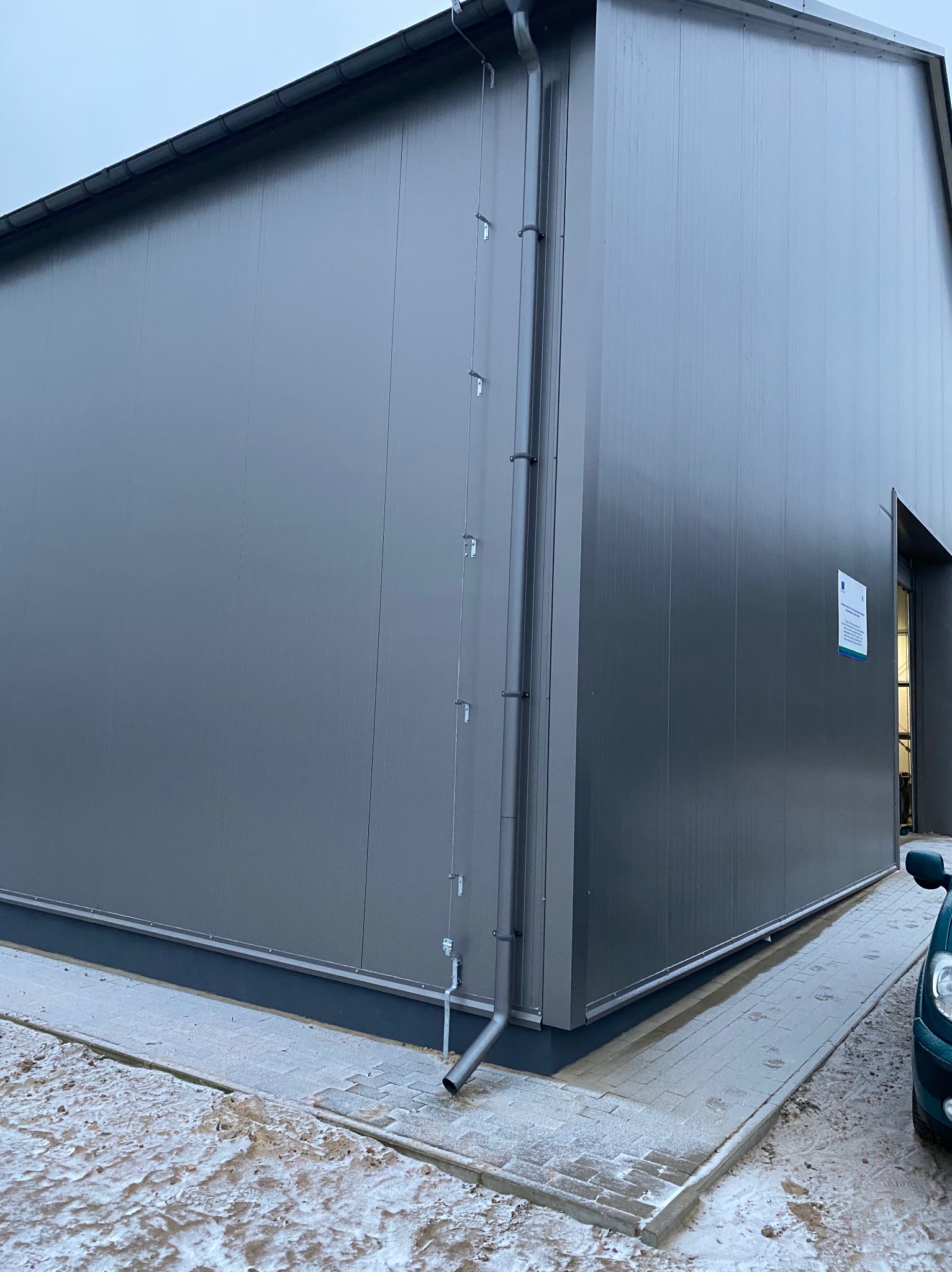 Szary budynek przemysłowy z blachy trapezowej, widoczny rynna spustowa, fragment chodnika i samochodu w zimowej scenerii.