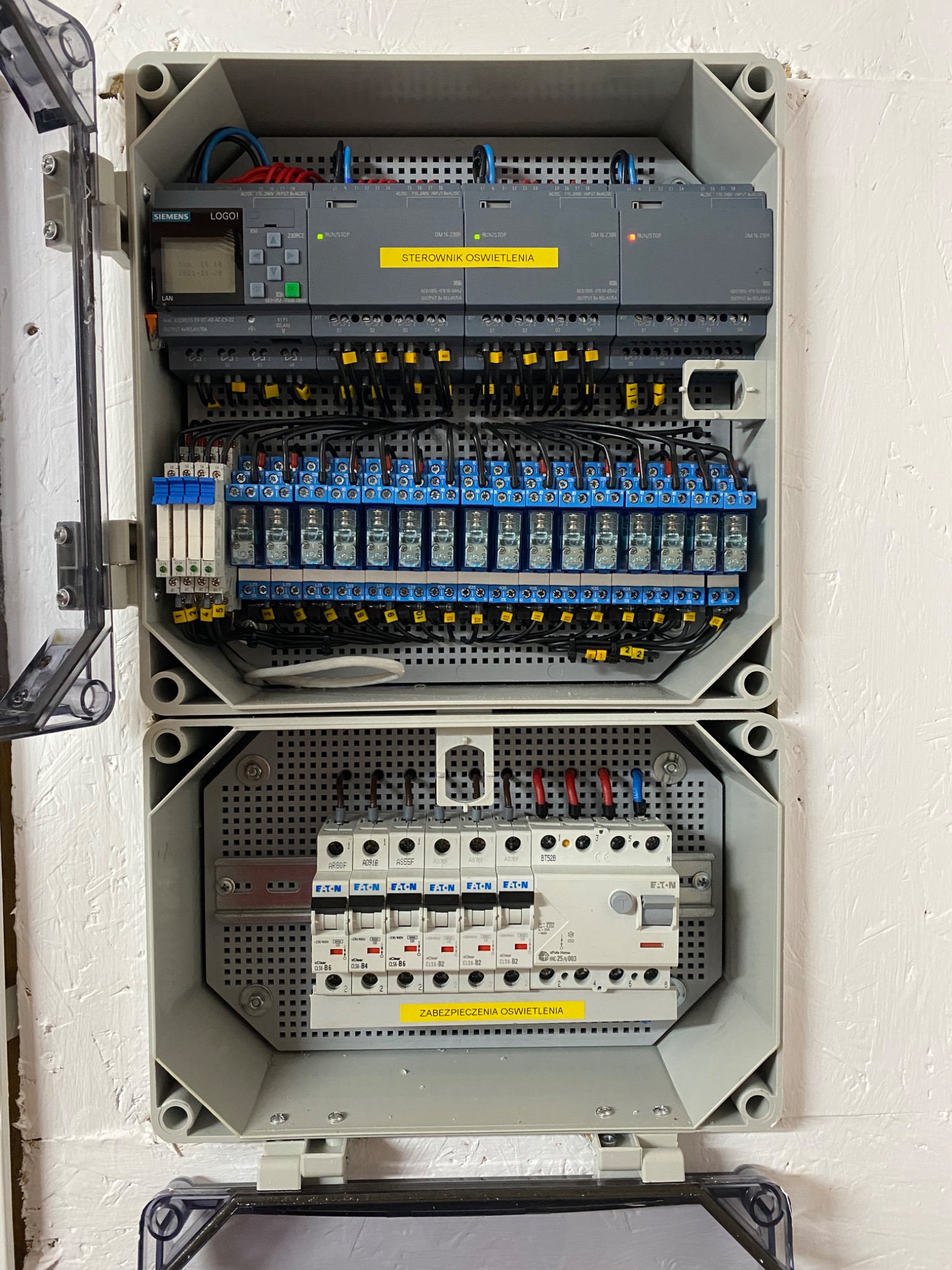 Otwarta szara szafka elektryczna z widocznymi elementami sterowania oświetleniem, w tym sterowniki Siemens LOGO!, przekaźniki i zabezpieczenia Eaton, zamontowana na białej ścianie o wyraźnej fakturze.