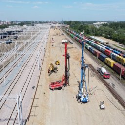 MENARD Sp. z o.o. - Szeroki kadr z lotu ptaka ukazuje plac budowy obok torów kolejowych, gdzie dwie palownice, czerwona i niebieska, pracują przy wzmacnianiu gruntu pod przyszłą infrastrukturę.