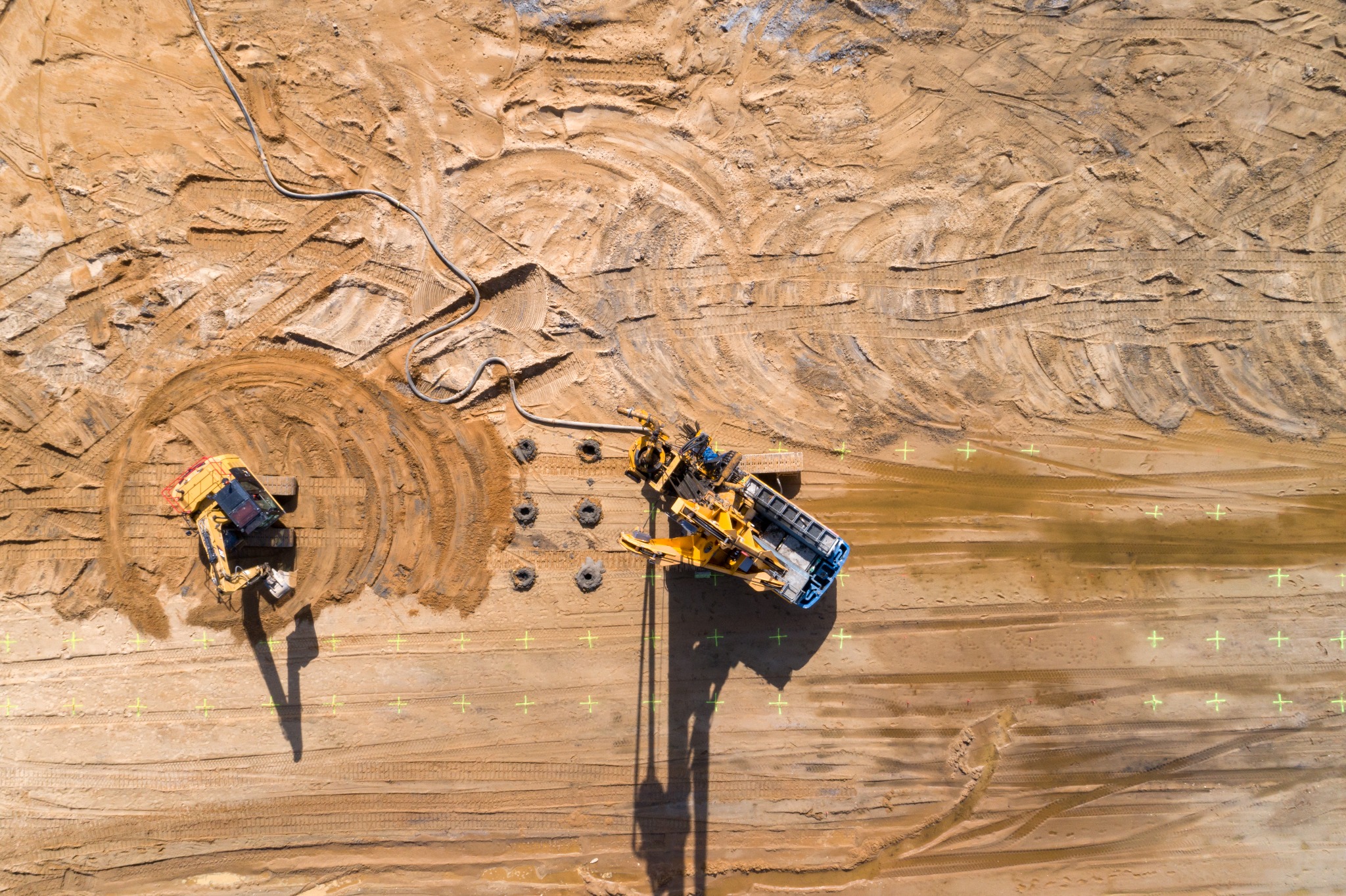 Zdjęcie z lotu ptaka przedstawia teren budowy z żółtym sprzętem do wiercenia otworów w ziemi i koparką, widoczne ślady opon i wyznaczone punkty pomiarowe na piaszczystym podłożu.