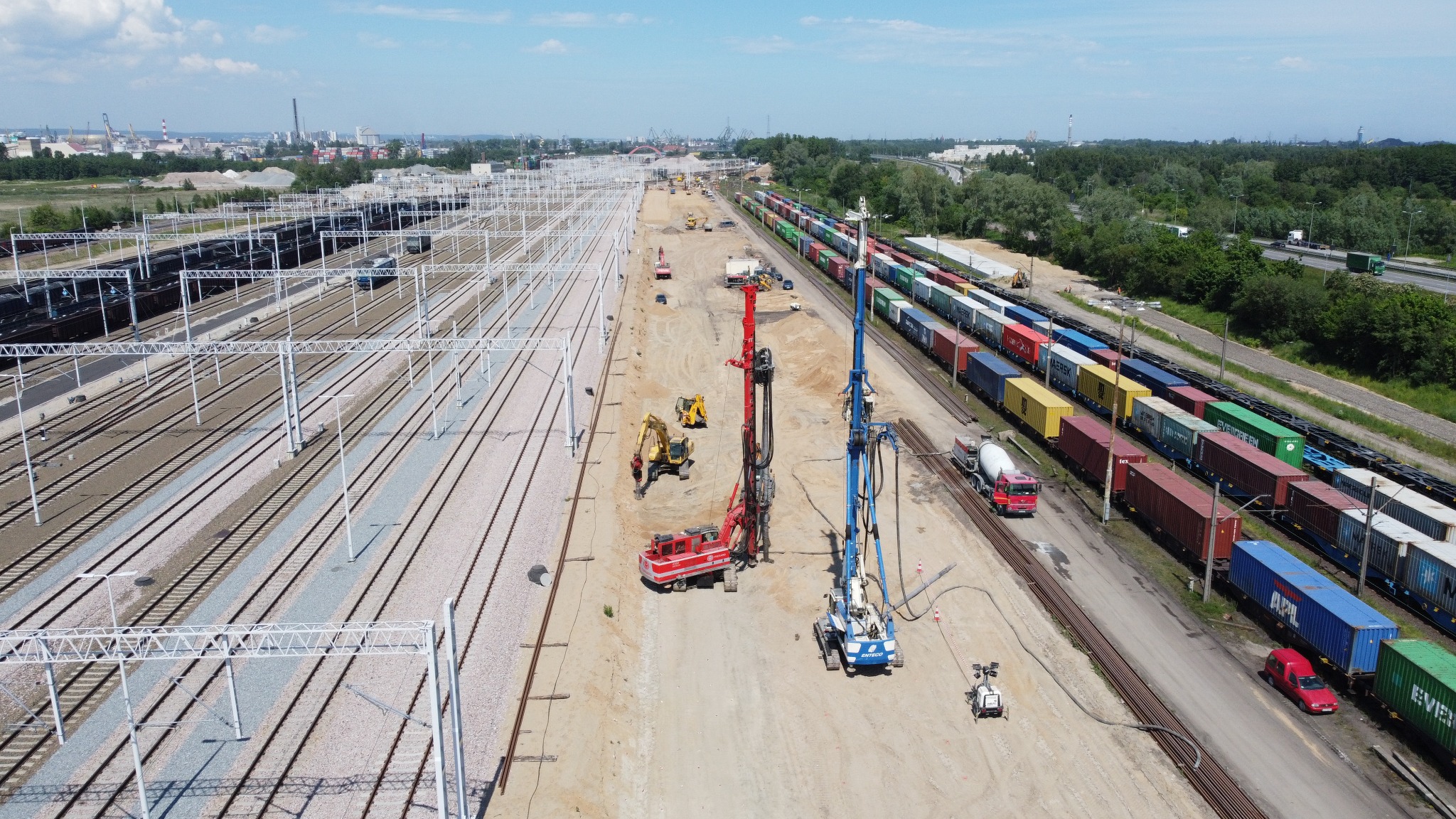 Szeroki kadr z lotu ptaka ukazuje plac budowy obok torów kolejowych, gdzie dwie palownice, czerwona i niebieska, pracują przy wzmacnianiu gruntu pod przyszłą infrastrukturę.