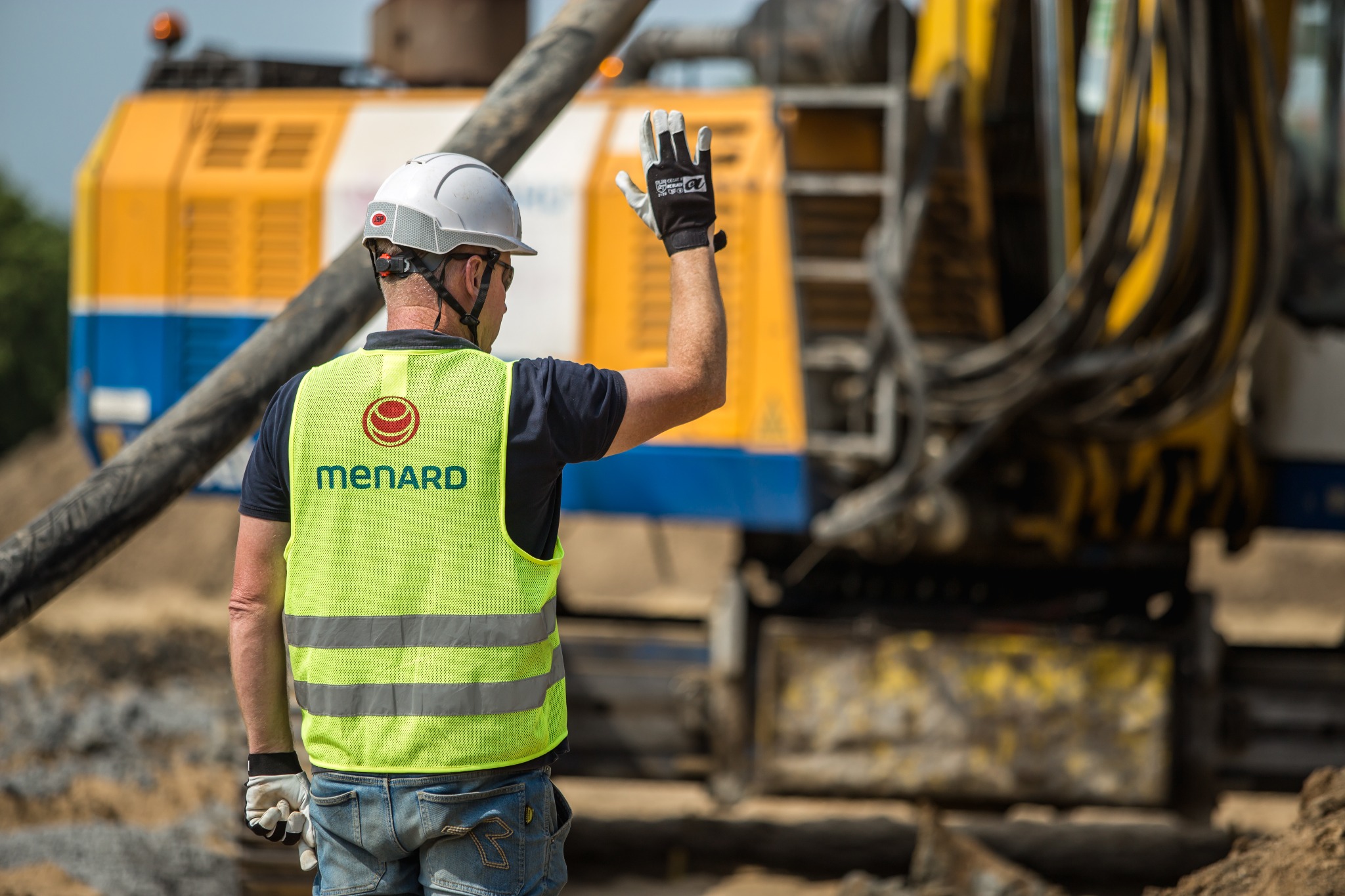 Pracownik w kasku i kamizelce odblaskowej Menard, sygnalizuje ręką na tle żółto-niebieskiej maszyny budowlanej i betonowej rury.