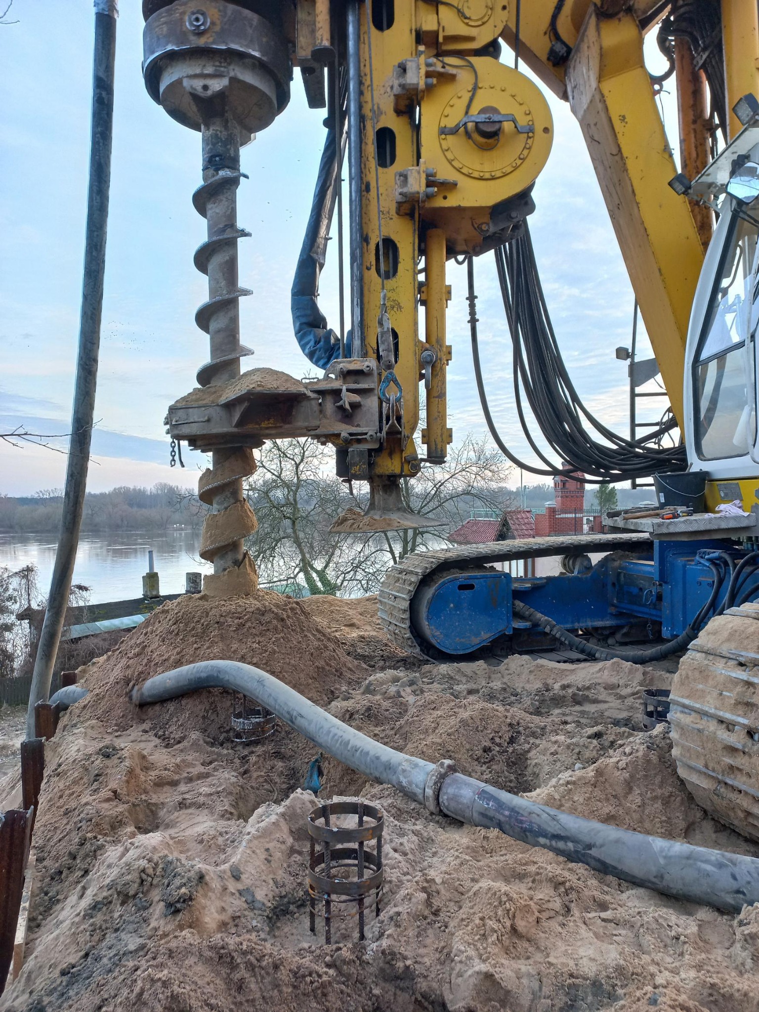 Żółta wiertnica na gąsienicach w trakcie wiercenia otworu w piaszczystym gruncie, widoczne stalowe zbrojenia i rura doprowadzająca płyn, w tle rzeka i drzewa.