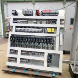 Szafa sterownicza z licznymi przekaźnikami, bezpiecznikami i okablowaniem, umieszczona na drewnianej palecie w hali produkcyjnej. Widoczny panel sterowania z przyciskiem alarmowym i ekranem.