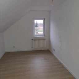 OLTECH Dawid Olek - Poddasze z oknem, grzejnikiem i podłogą panelową; widoczne gniazdka elektryczne i wisząca oprawa oświetleniowa bez klosza.