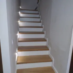 Schody drewniane z białymi podstopnicami i podświetleniem LED wzdłuż ściany, widok od dołu.