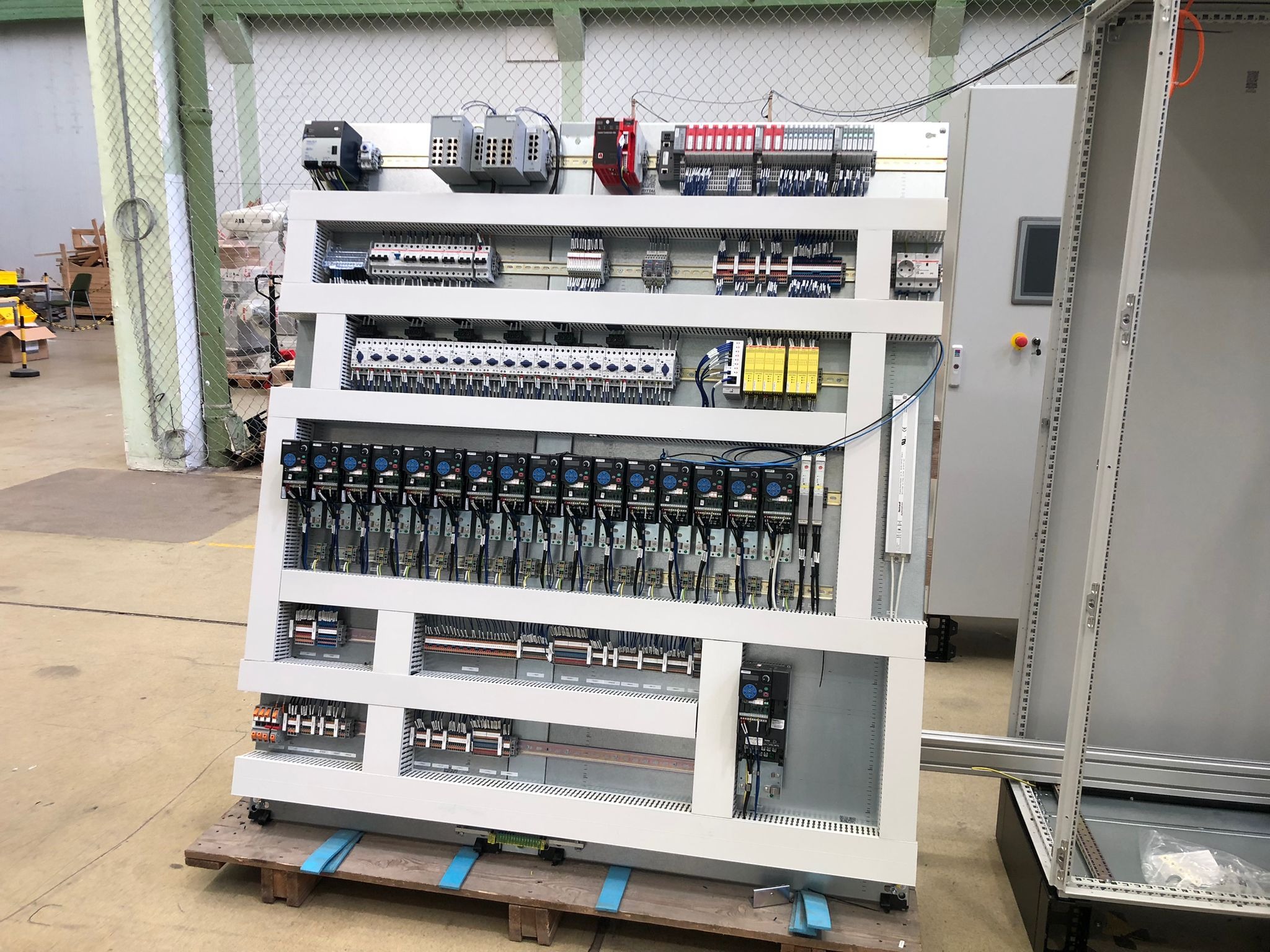 Szafa sterownicza z licznymi przekaźnikami, bezpiecznikami i okablowaniem, umieszczona na drewnianej palecie w hali produkcyjnej. Widoczny panel sterowania z przyciskiem alarmowym i ekranem.