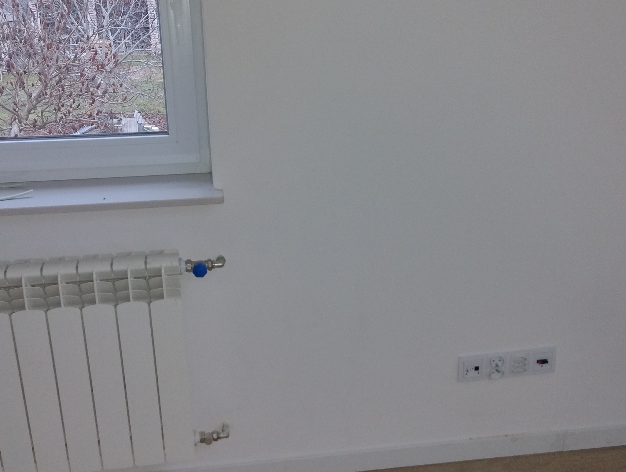 Biała ściana z grzejnikiem z niebieskim zaworem, gniazdkami elektrycznymi i widokiem z okna na drzewo bez liści.