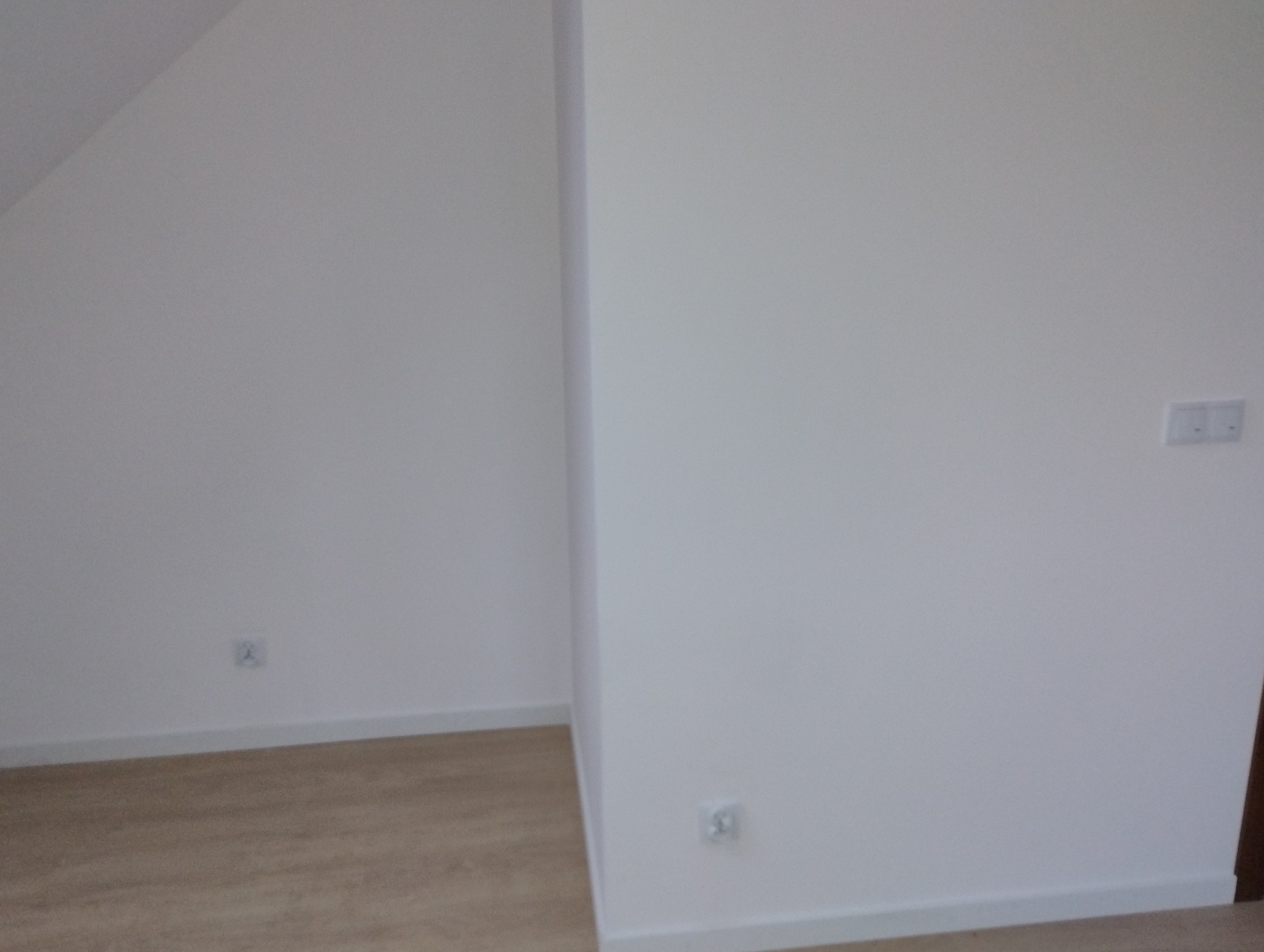 Pusty, świeżo wykończony pokój z jasnymi ścianami, widoczne gniazdka elektryczne na ścianach i włącznik światła, podłoga laminowana imitująca drewno, listwy przypodłogowe.