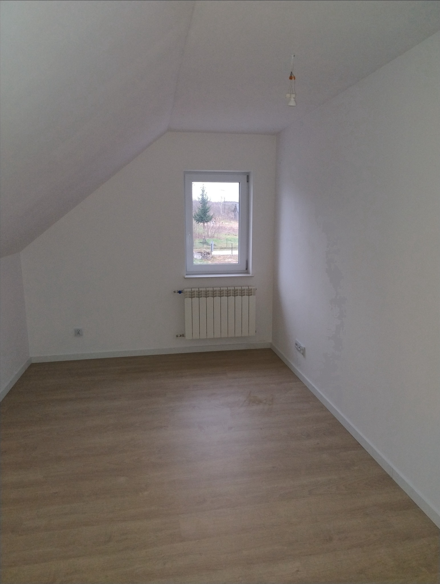 Poddasze z oknem, grzejnikiem i podłogą panelową; widoczne gniazdka elektryczne i wisząca oprawa oświetleniowa bez klosza.