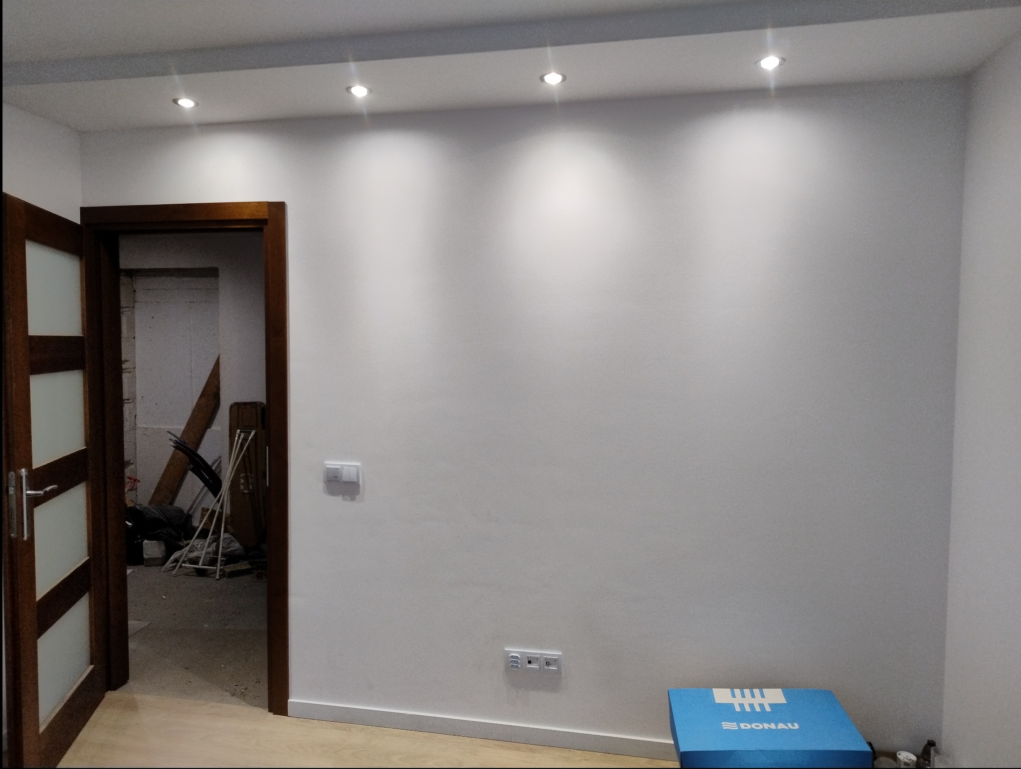 Białe wnętrze z punktowym oświetleniem sufitowym, widoczne gniazdka elektryczne oraz otwarte drzwi do pomieszczenia w trakcie remontu.
