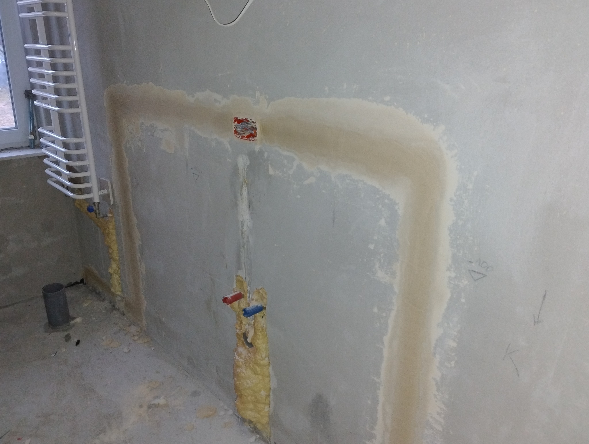 Ściana w trakcie remontu z widocznym podłączeniem grzejnika, instalacją elektryczną w puszce oraz zakrytymi pianką rurami.