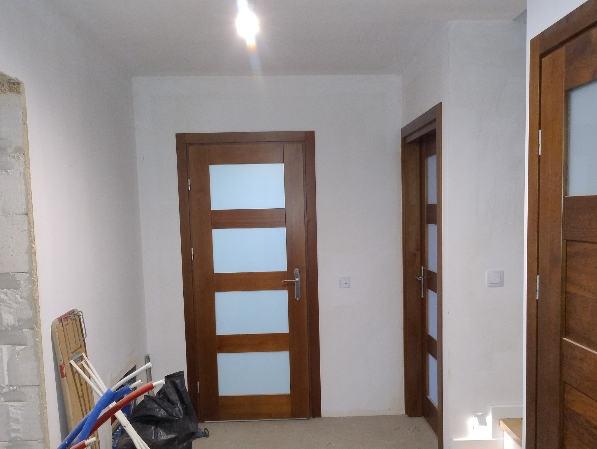 Dwa drewniane drzwi wewnętrzne z szybami mlecznymi w nowym, jeszcze nieumeblowanym pomieszczeniu. Widoczne przewody i materiały budowlane sugerują prace wykończeniowe.