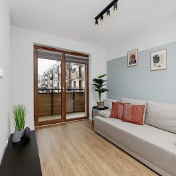 Wiktoria Rola - Jasne wnętrze mieszkania we Wrocławiu z balkonem, widok na osiedle, drewniane panele, sofa z pomarańczowymi poduszkami, roślina doniczkowa obok balkonu.