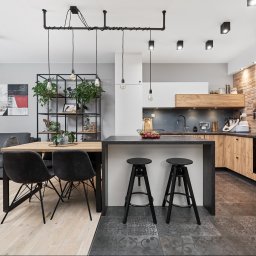 Wiktoria Rola - Jasne, przestronne wnętrze mieszkania z otwartą kuchnią, drewnianym stołem, czarnymi krzesłami i wyspą kuchenną z hokerami. W tle szara sofa i abstrakcyjny obraz.