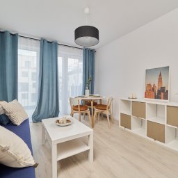 Wiktoria Rola - Jasne, nowocześnie urządzone wnętrze mieszkania z niebieską sofą, białym stolikiem kawowym i okrągłym stołem jadalnym z krzesłami. Na ścianie wisi obraz z motywem miejskim.