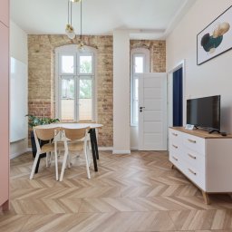 Wiktoria Rola - Jasne wnętrze mieszkania z parkietem w jodełkę, ceglaną ścianą, białymi oknami i nowoczesnymi meblami w pastelowych kolorach.