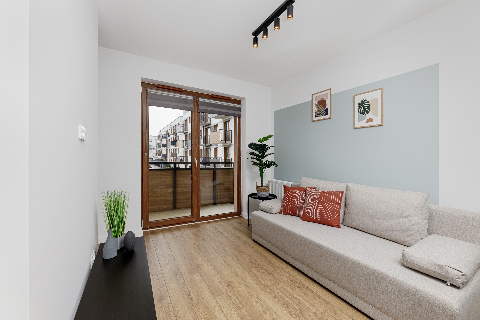 Jasne wnętrze mieszkania we Wrocławiu z balkonem, widok na osiedle, drewniane panele, sofa z pomarańczowymi poduszkami, roślina doniczkowa obok balkonu.