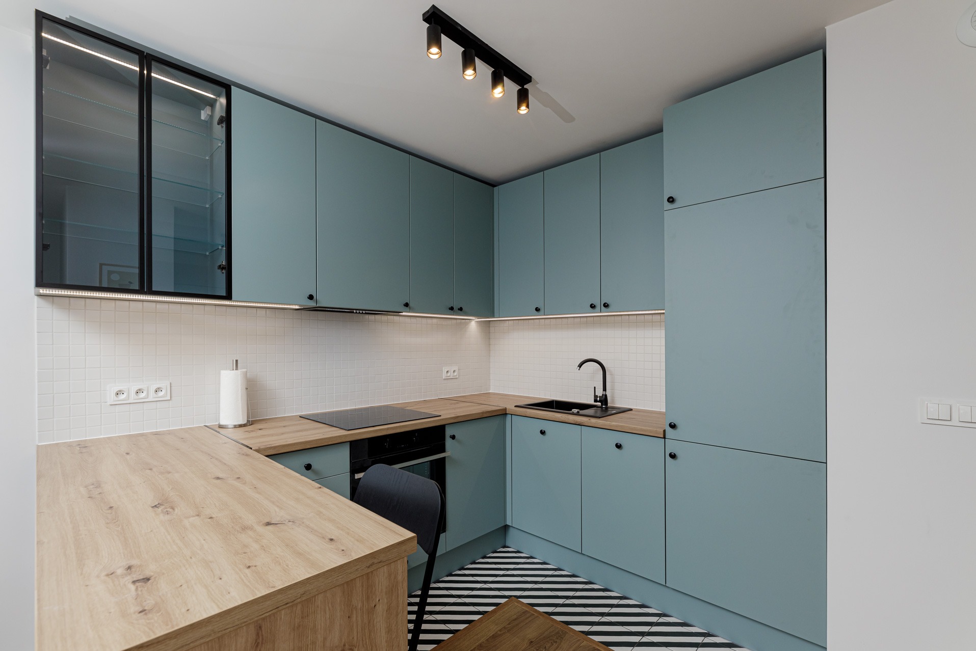 Nowoczesna kuchnia w odcieniach błękitu z drewnianymi blatami, czarnymi akcentami i wzorzystą podłogą w paski, oświetlona listwami LED pod szafkami górnymi.