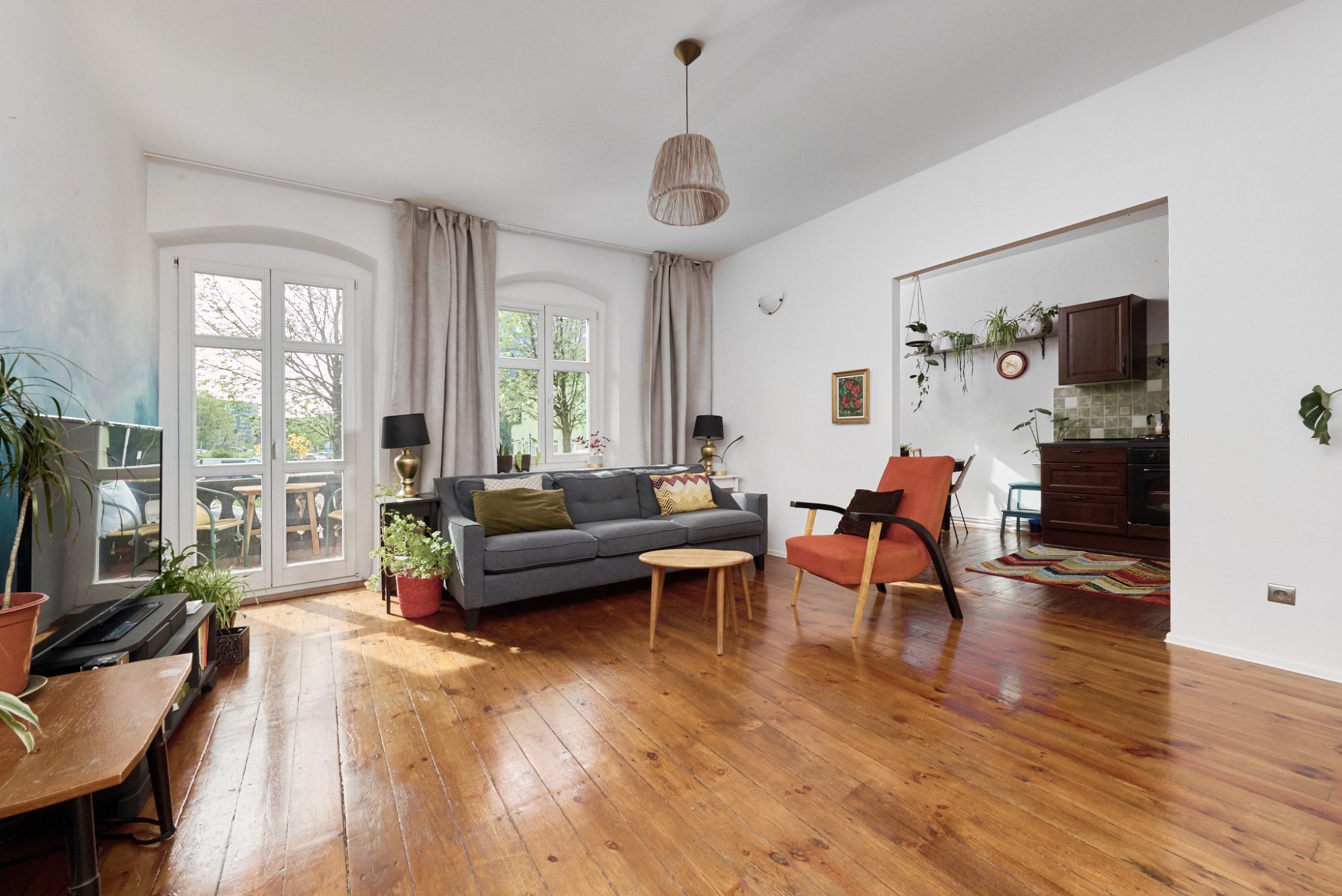 Jasne, przestronne wnętrze z drewnianą podłogą, szarą sofą, pomarańczowym fotelem i widokiem na balkon przez dwuskrzydłowe drzwi.