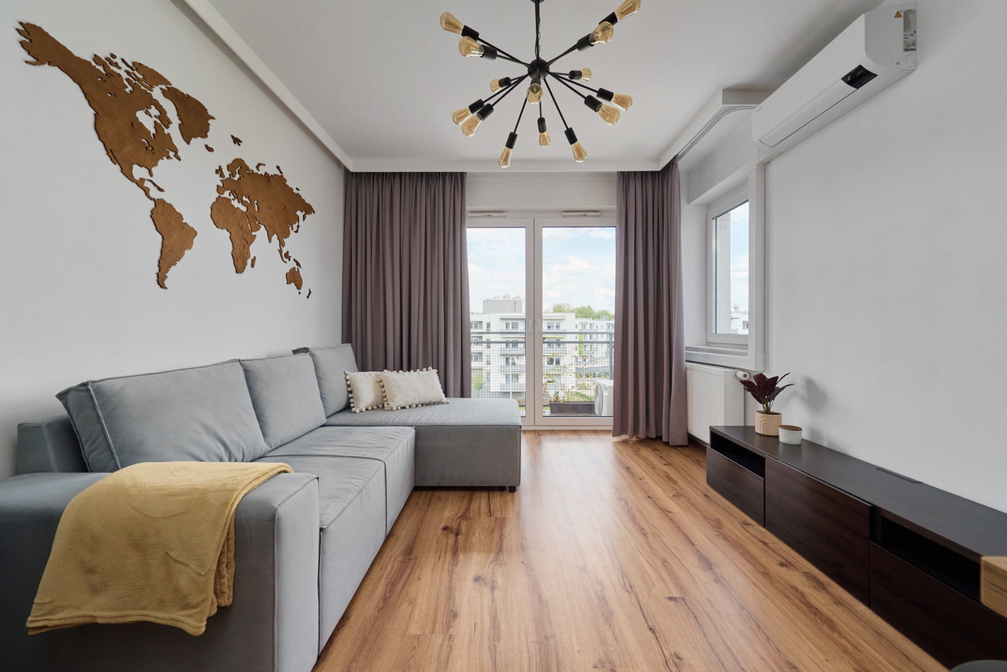 Jasne wnętrze salonu z szarym narożnikiem, drewnianą mapą świata na ścianie i parkietem, oświetlone lampą sufitową z żarówkami Edison.