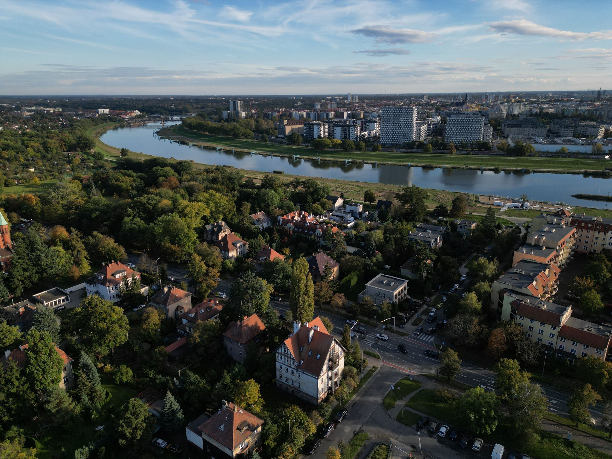 Panoramiczny widok z lotu ptaka na miasto z rzeką, zabudową mieszkaniową i zielenią, w słoneczny dzień.