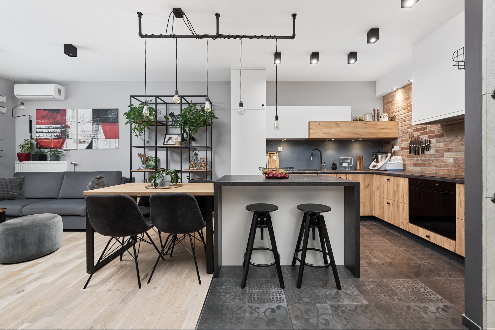 Jasne, przestronne wnętrze mieszkania z otwartą kuchnią, drewnianym stołem, czarnymi krzesłami i wyspą kuchenną z hokerami. W tle szara sofa i abstrakcyjny obraz.