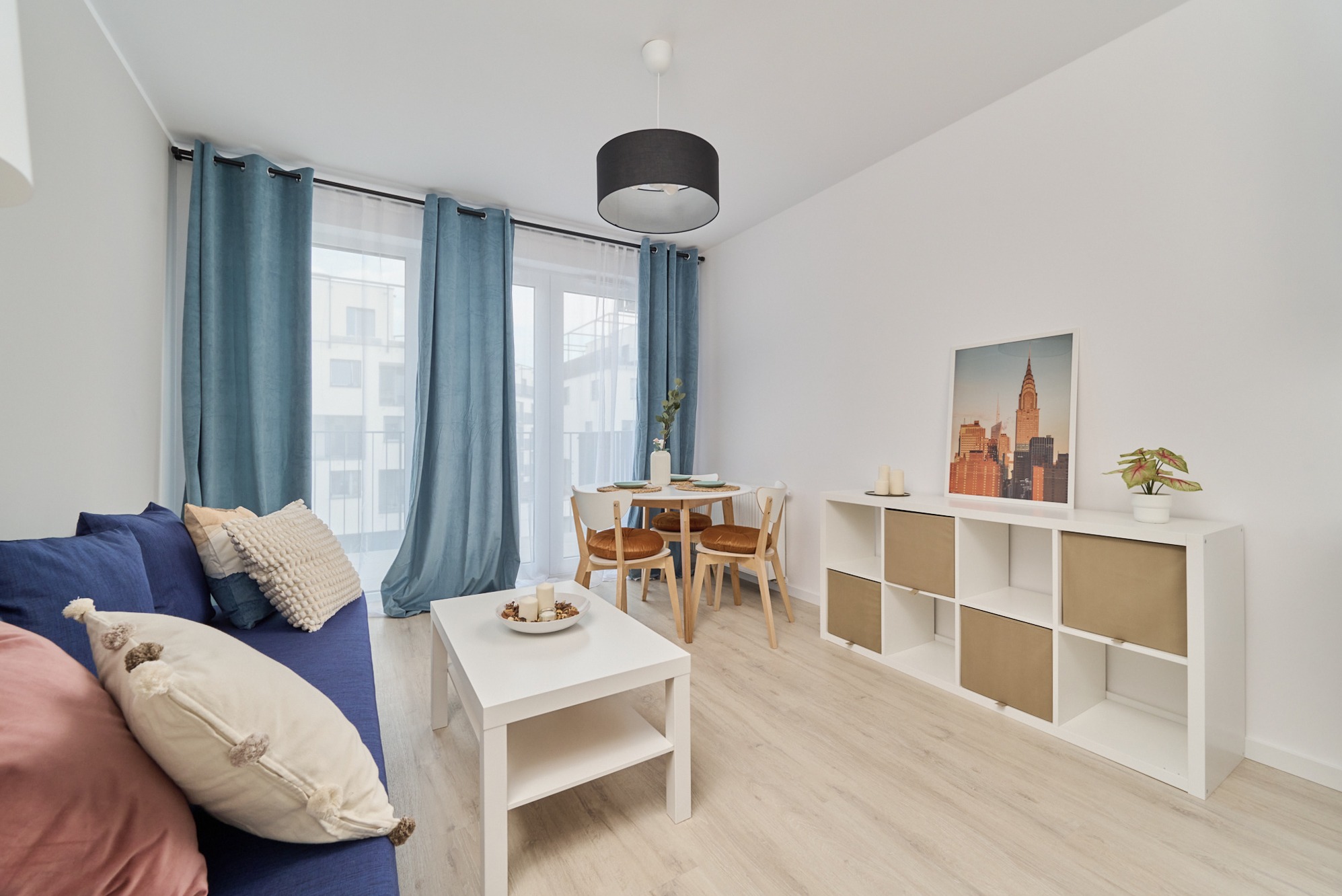 Jasne, nowocześnie urządzone wnętrze mieszkania z niebieską sofą, białym stolikiem kawowym i okrągłym stołem jadalnym z krzesłami. Na ścianie wisi obraz z motywem miejskim.