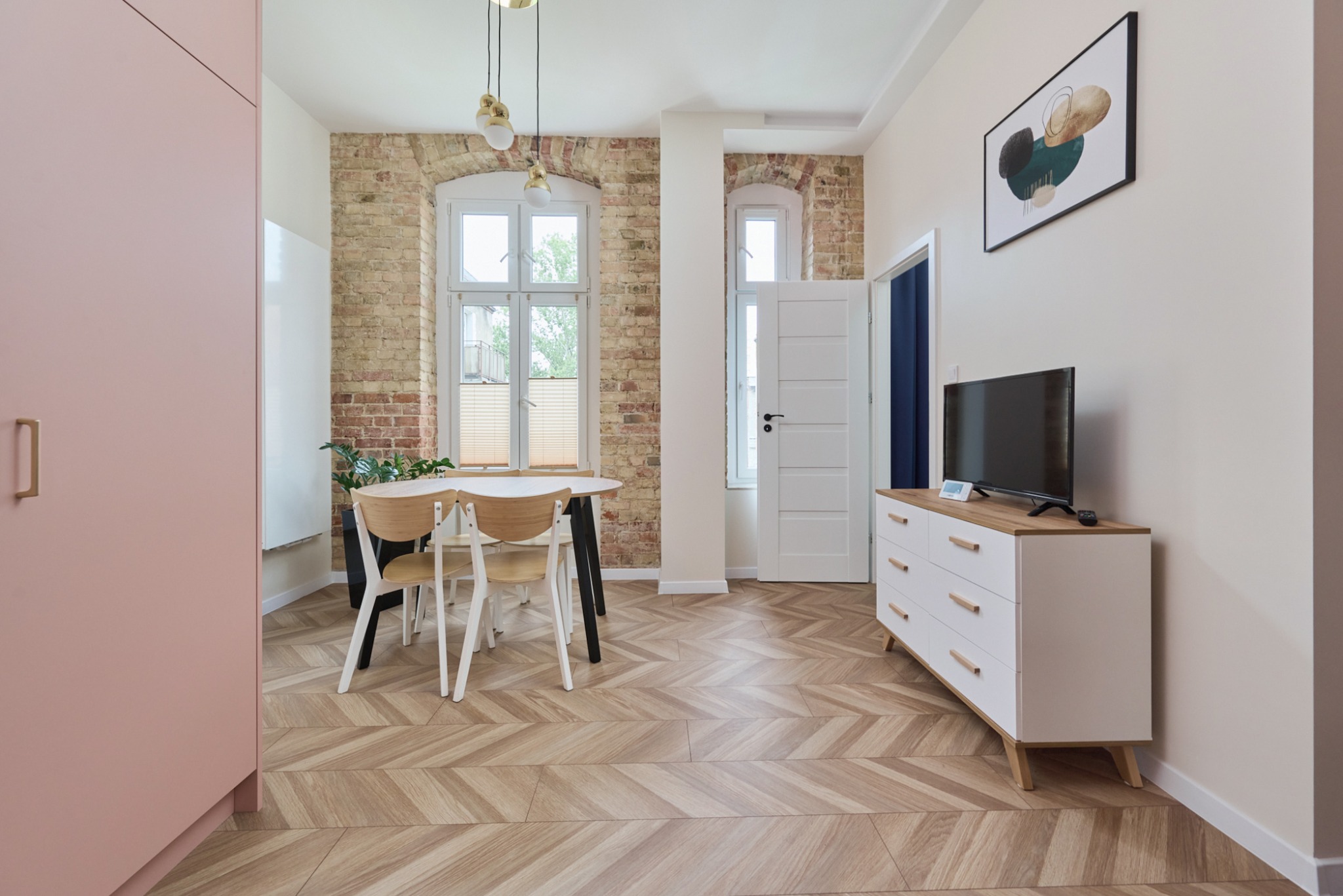 Jasne wnętrze mieszkania z parkietem w jodełkę, ceglaną ścianą, białymi oknami i nowoczesnymi meblami w pastelowych kolorach.