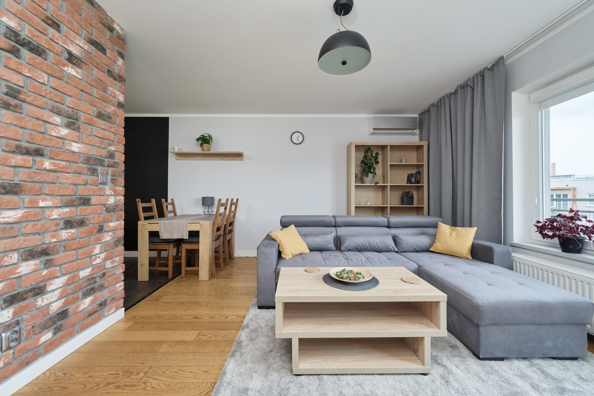 Jasne, przestronne wnętrze salonu z ceglaną ścianą, drewnianą podłogą, szarą sofą narożną i drewnianym stołem kawowym. W tle widoczny stół jadalny i krzesła.