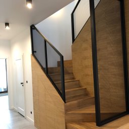 spawaniewarszawa.pl Karolina Szydłowska - Nowoczesne schody z drewnianymi stopniami i czarną metalową balustradą z perforowanym wypełnieniem, zintegrowane z zabudową meblową pod schodami, oświetlone punktowymi lampami sufitowymi.