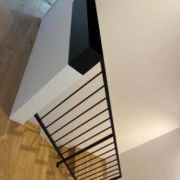 spawaniewarszawa.pl Karolina Szydłowska - Minimalistyczne schody z czarnymi, metalowymi poręczami i drewnianymi stopniami widziane z góry, z fragmentem białej ściany i czarną obudową.