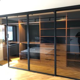 spawaniewarszawa.pl Karolina Szydłowska - Szklane drzwi przesuwne z czarnymi metalowymi ramami oddzielające garderobę z drewnianymi półkami i komodą, widok z parkietu w jodełkę.