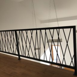 spawaniewarszawa.pl Karolina Szydłowska - Nowoczesna balustrada wewnętrzna z czarnych, metalowych prętów tworzących geometryczny wzór krzyży, zamontowana na drewnianej podłodze, z widocznymi lampami wiszącymi i oknem w tle.