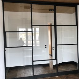 spawaniewarszawa.pl Karolina Szydłowska - Nowoczesne drzwi szklane z czarnymi metalowymi ramami, widok na parkiet w jodełkę i białe ściany, z odbiciem sylwetki osoby w szybie.