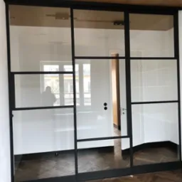 Nowoczesne drzwi szklane z czarnymi metalowymi ramami, widok na parkiet w jodełkę i białe ściany, z odbiciem sylwetki osoby w szybie.