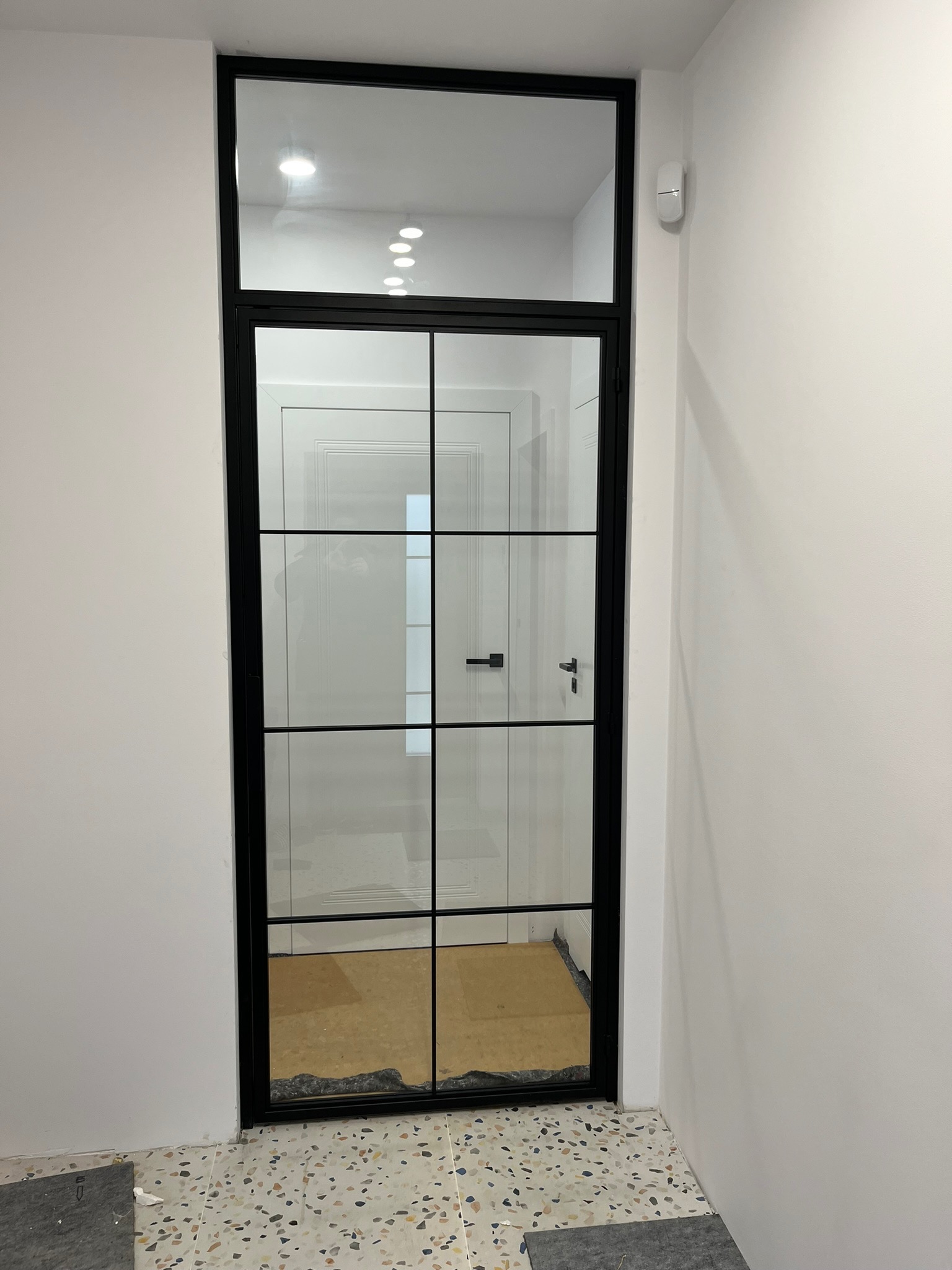 Nowoczesne drzwi szklane z czarnymi szprosami, widok na białe wnętrze z oświetleniem punktowym, posadzka lastryko.