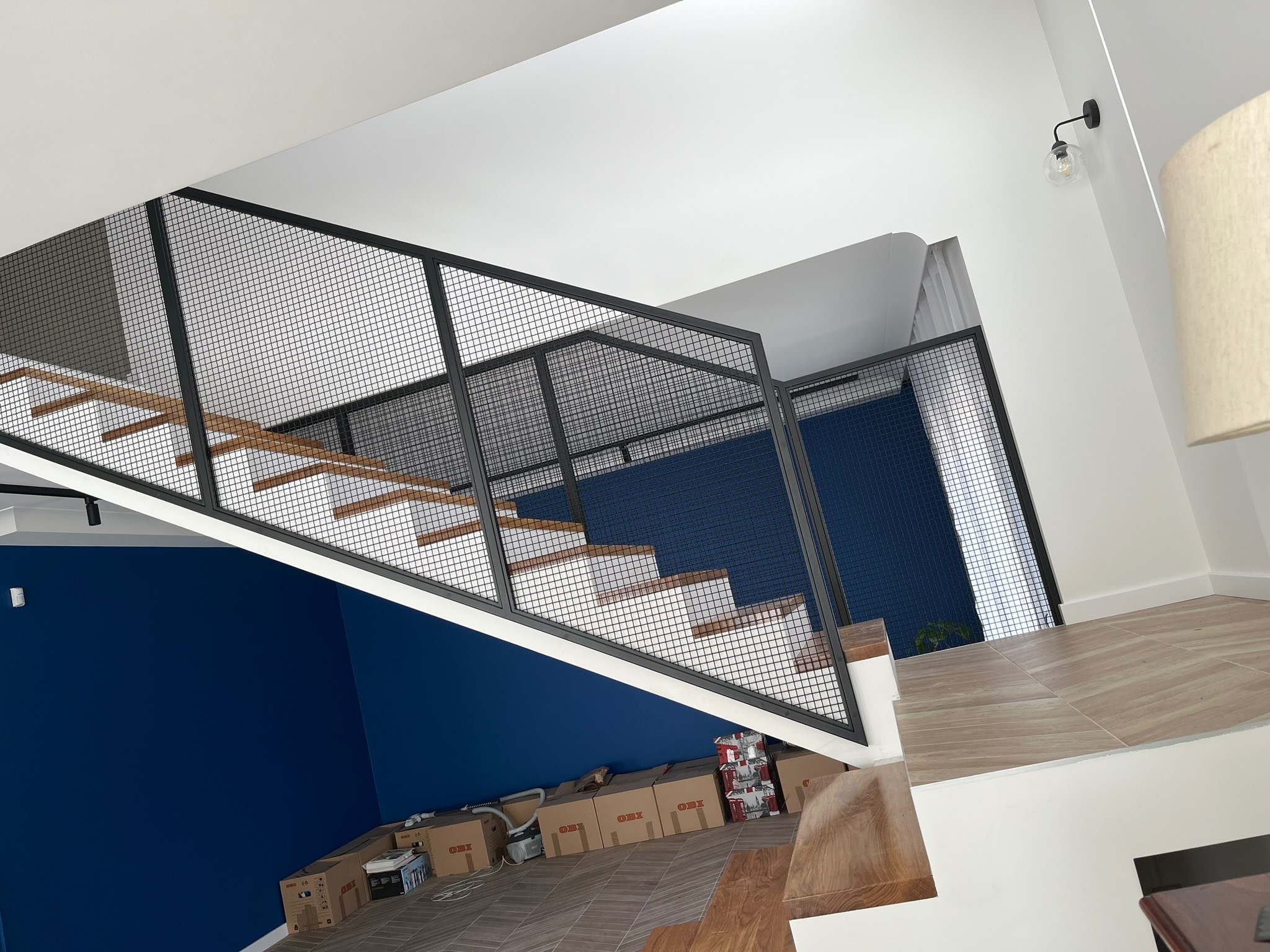 Nowoczesne schody z drewnianymi stopniami i czarną metalową ażurową balustradą, widok z góry na niebieską ścianę i kartony na podłodze.