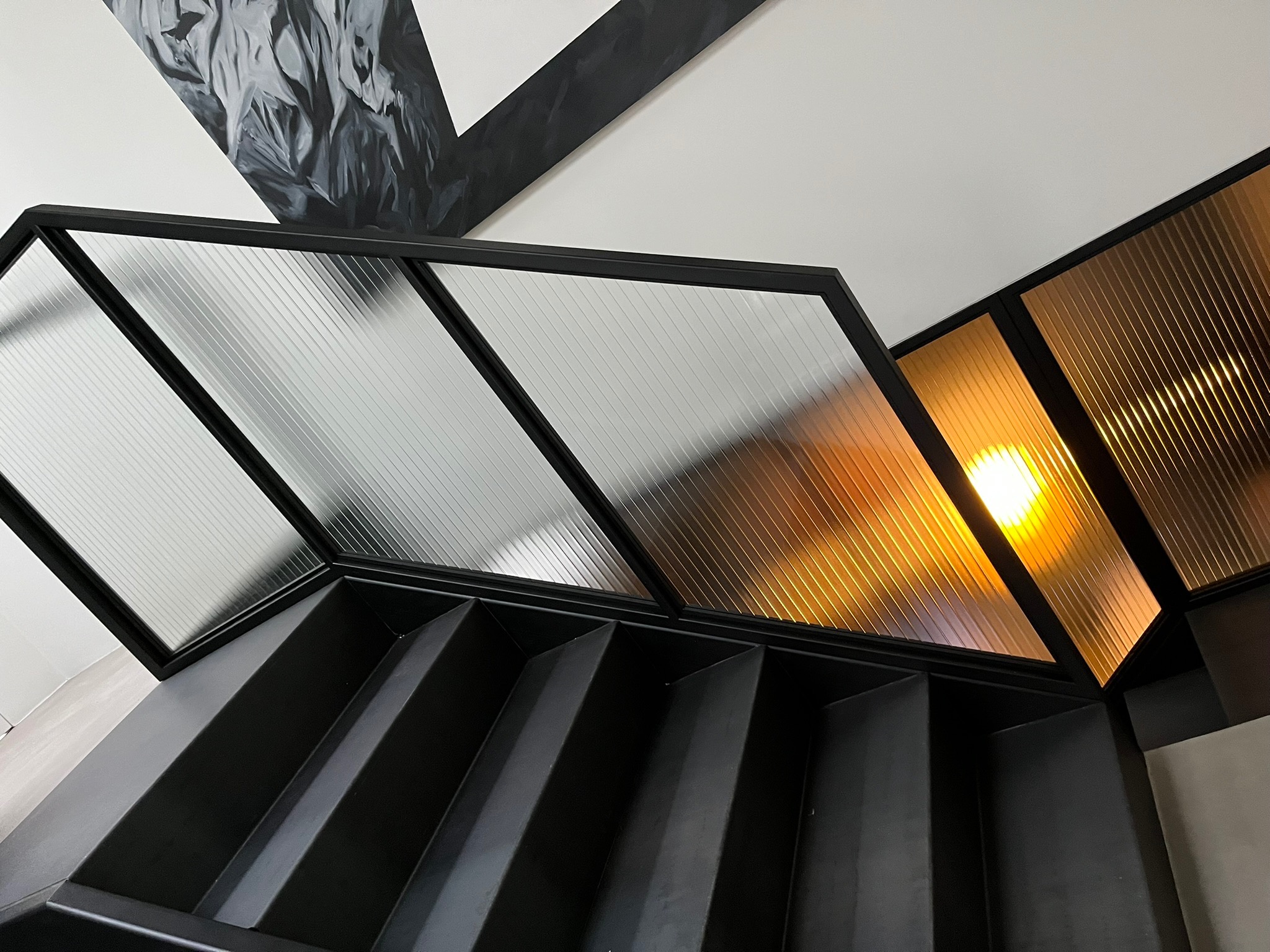Nowoczesne, czarne schody z przeszklonym, geometrycznym zadaszeniem, częściowo z przezroczystego i częściowo z bursztynowego szkła, oświetlone od spodu, z abstrakcyjną dekoracją na ścianie.