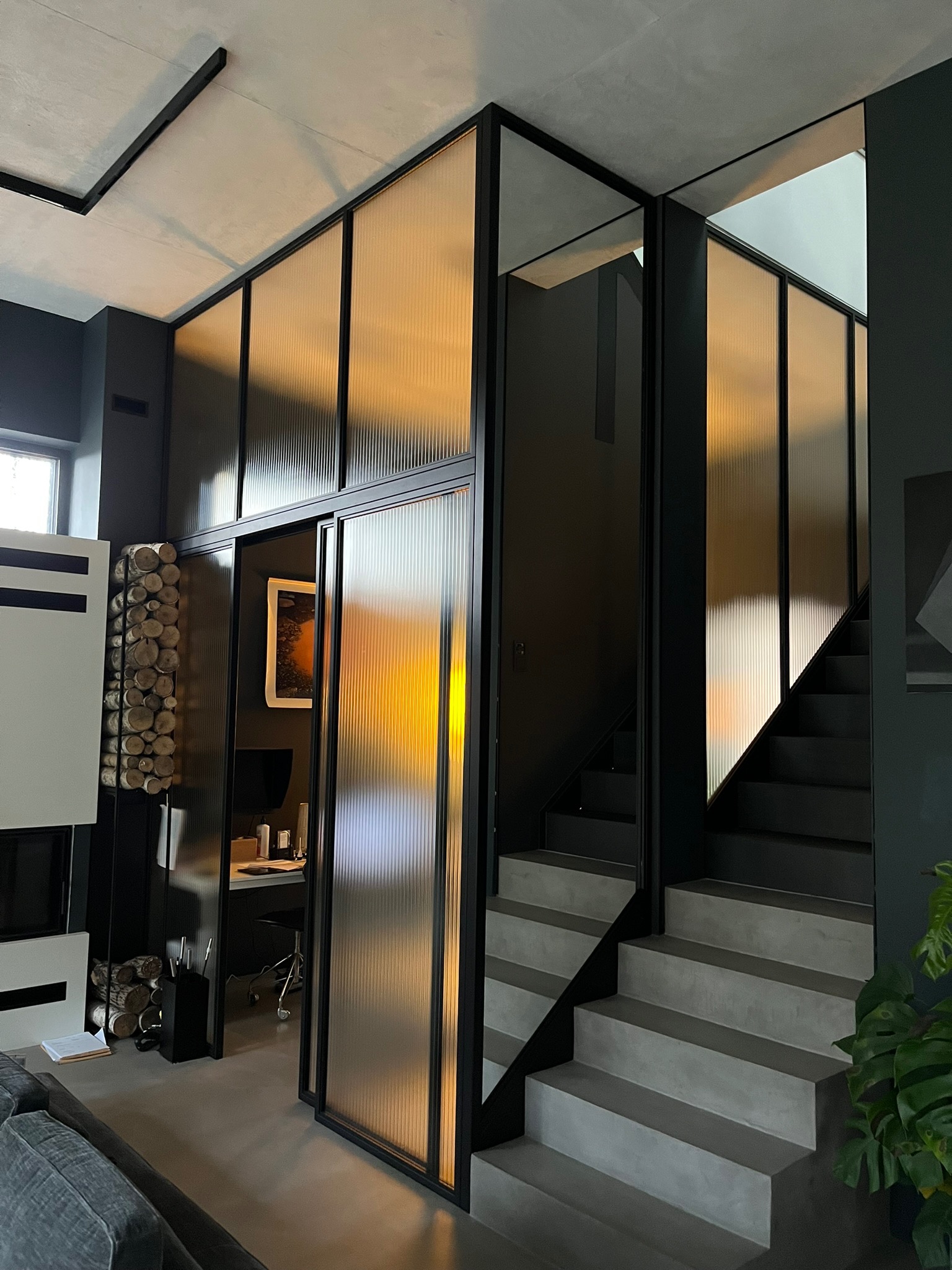 Nowoczesne wnętrze z szarymi betonowymi schodami, przeszkloną ścianą z czarnymi ramami i widokiem na biuro za przesuwnymi drzwiami.