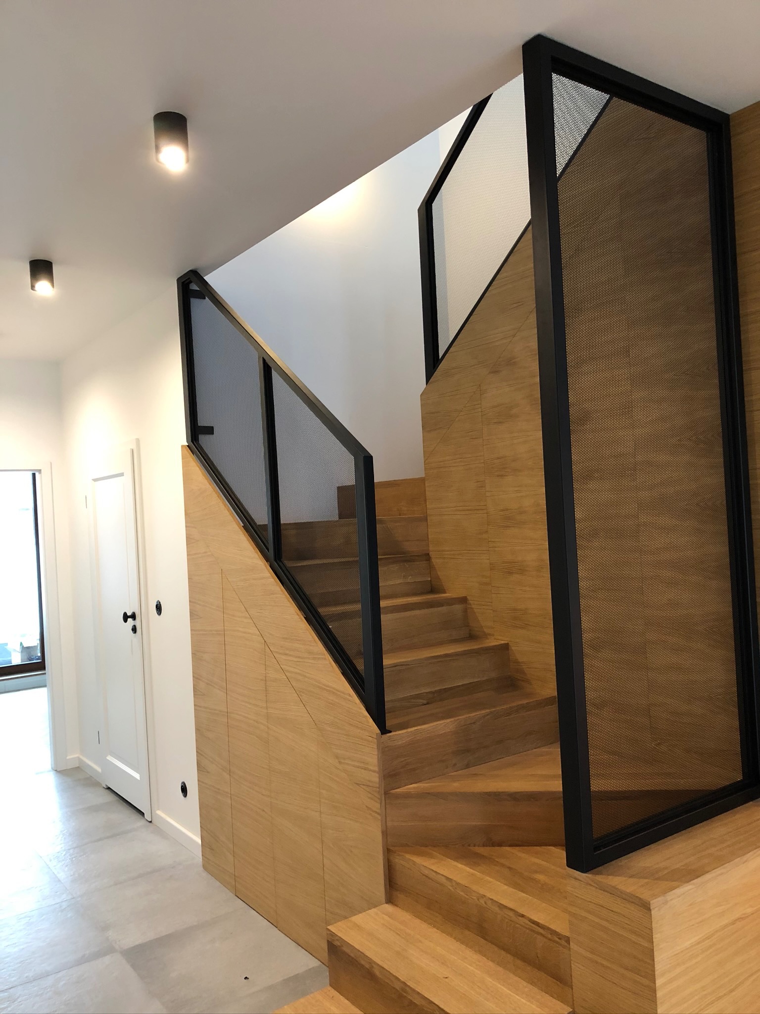 Nowoczesne schody z drewnianymi stopniami i czarną metalową balustradą z perforowanym wypełnieniem, zintegrowane z zabudową meblową pod schodami, oświetlone punktowymi lampami sufitowymi.
