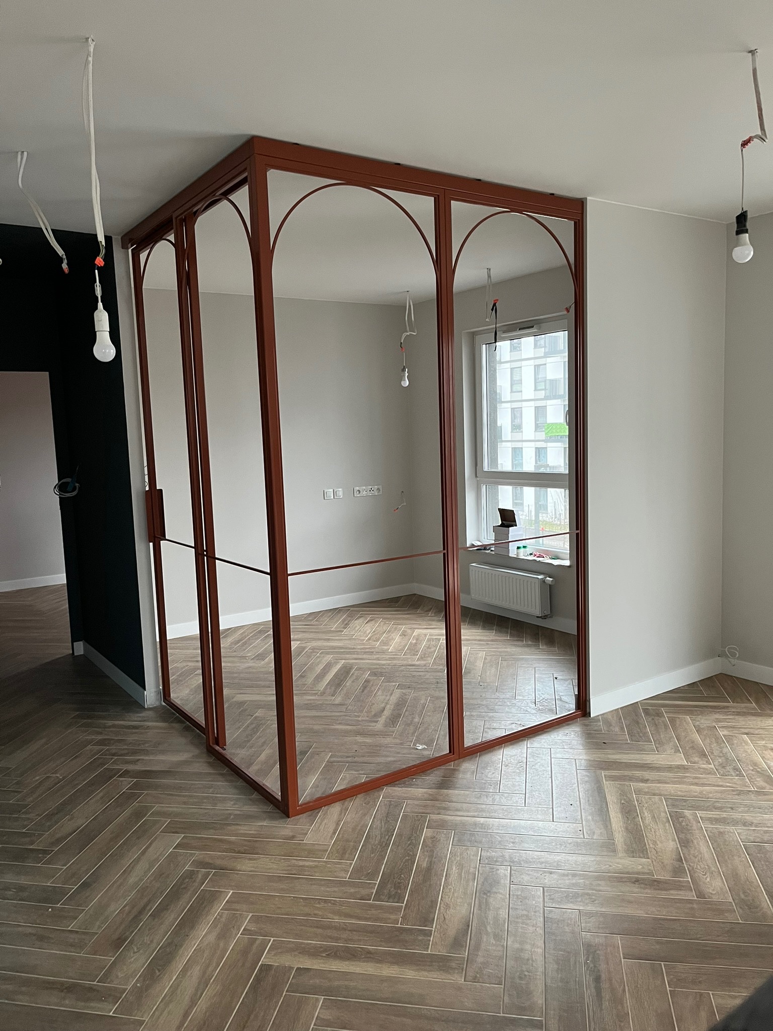 Nowoczesna, brązowa, metalowa konstrukcja z łukami i szklanymi panelami, prawdopodobnie drzwi szklane lub przegroda, w surowym wnętrzu z parkietem ułożonym w jodełkę i wiszącymi kablami...