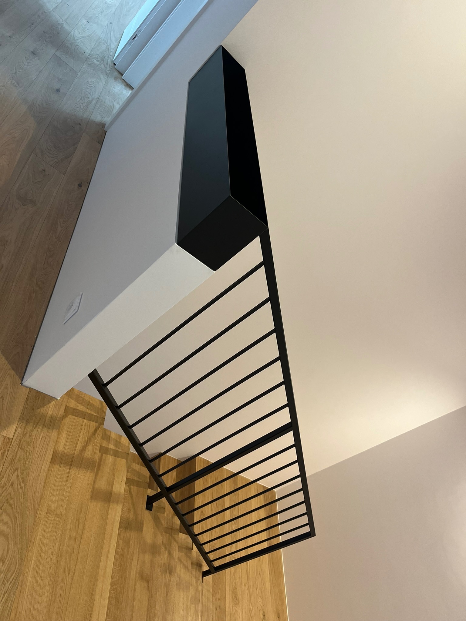 Minimalistyczne schody z czarnymi, metalowymi poręczami i drewnianymi stopniami widziane z góry, z fragmentem białej ściany i czarną obudową.