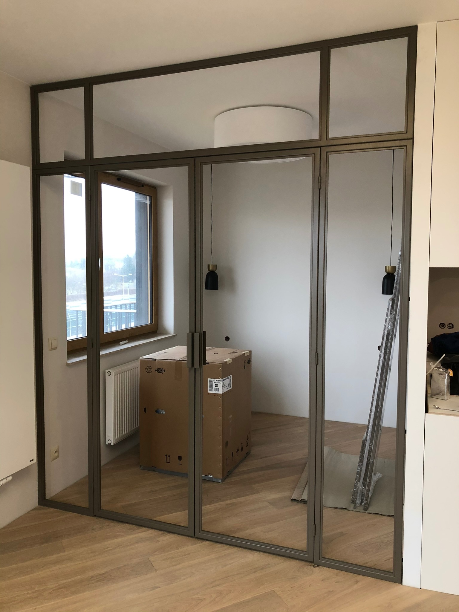 Szklane drzwi z metalową ramą w stylu loft, widoczne kartony i drabina w tle, podłoga z paneli, wnętrze w trakcie wykańczania.