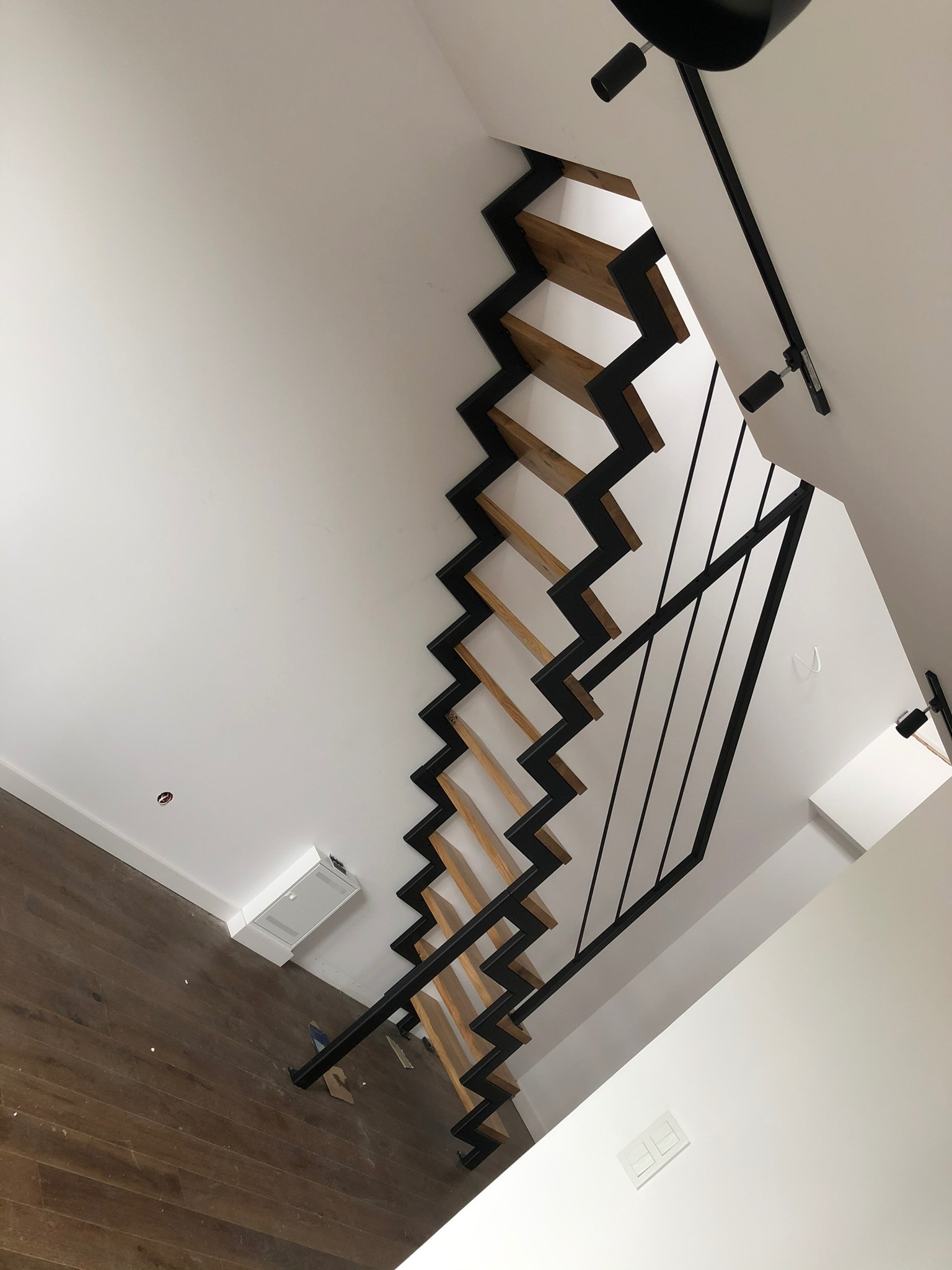 Nowoczesne schody z drewnianymi stopniami i czarną metalową konstrukcją, widok od dołu, oświetlenie punktowe na suficie.