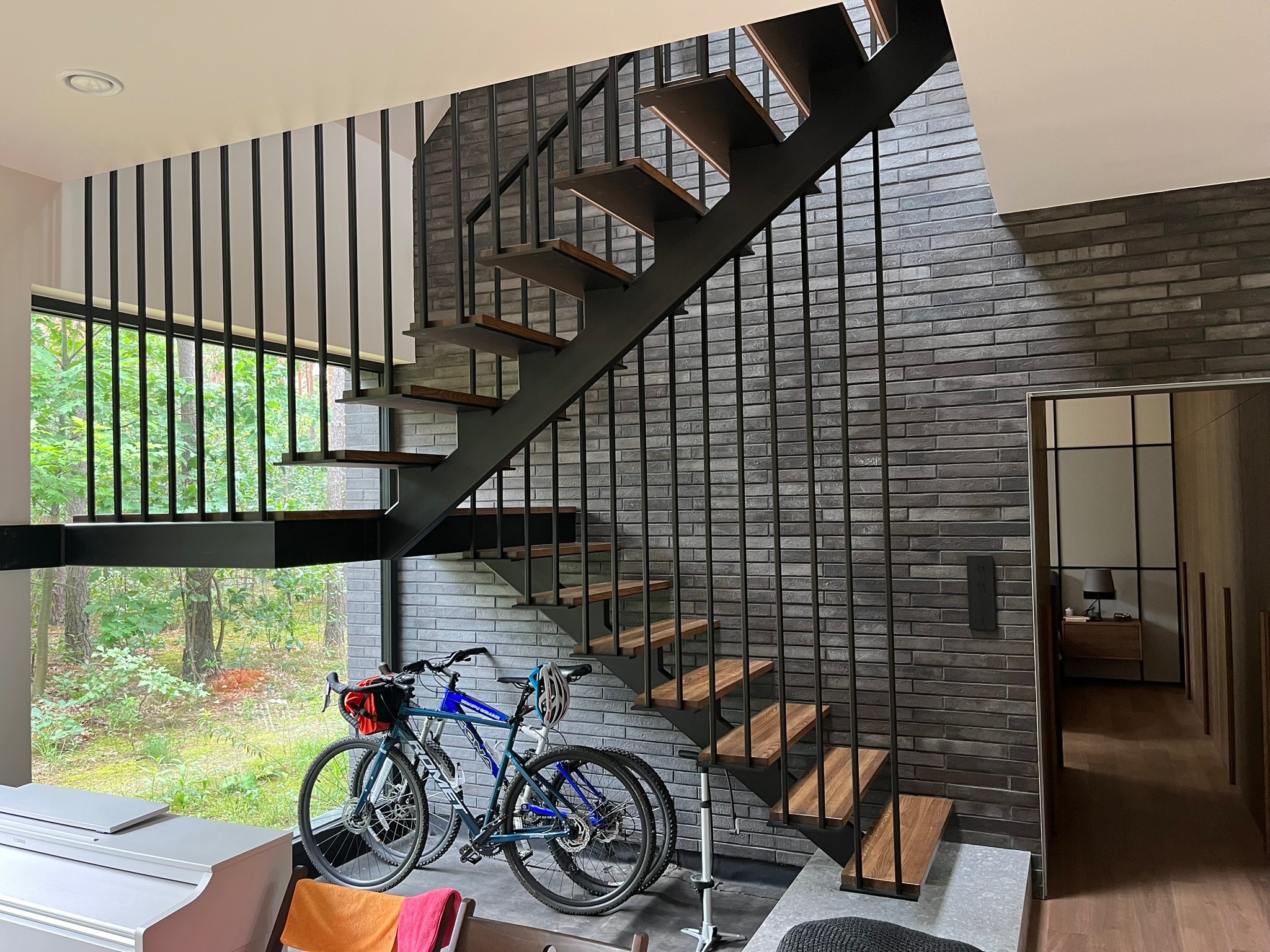 Nowoczesne schody z metalową konstrukcją i drewnianymi stopniami, widoczne na tle ściany z szarej cegły, z rowerami zaparkowanymi u podnóża schodów i widokiem na zieleń za oknem.
