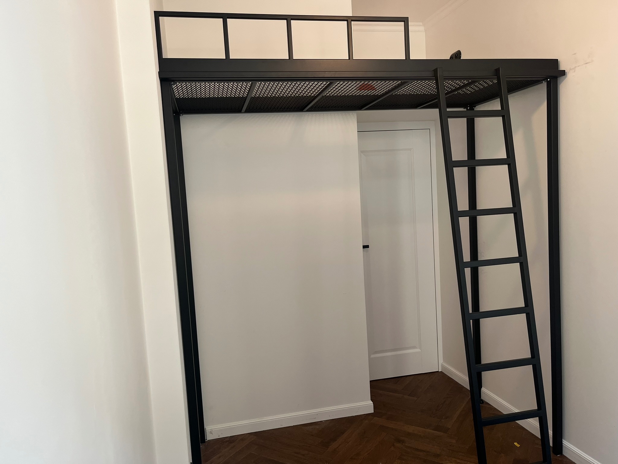 Czarna metalowa konstrukcja łóżka na antresoli z drabinką, widoczna na tle białej ściany i drzwi, z parkietem w jodełkę na podłodze.